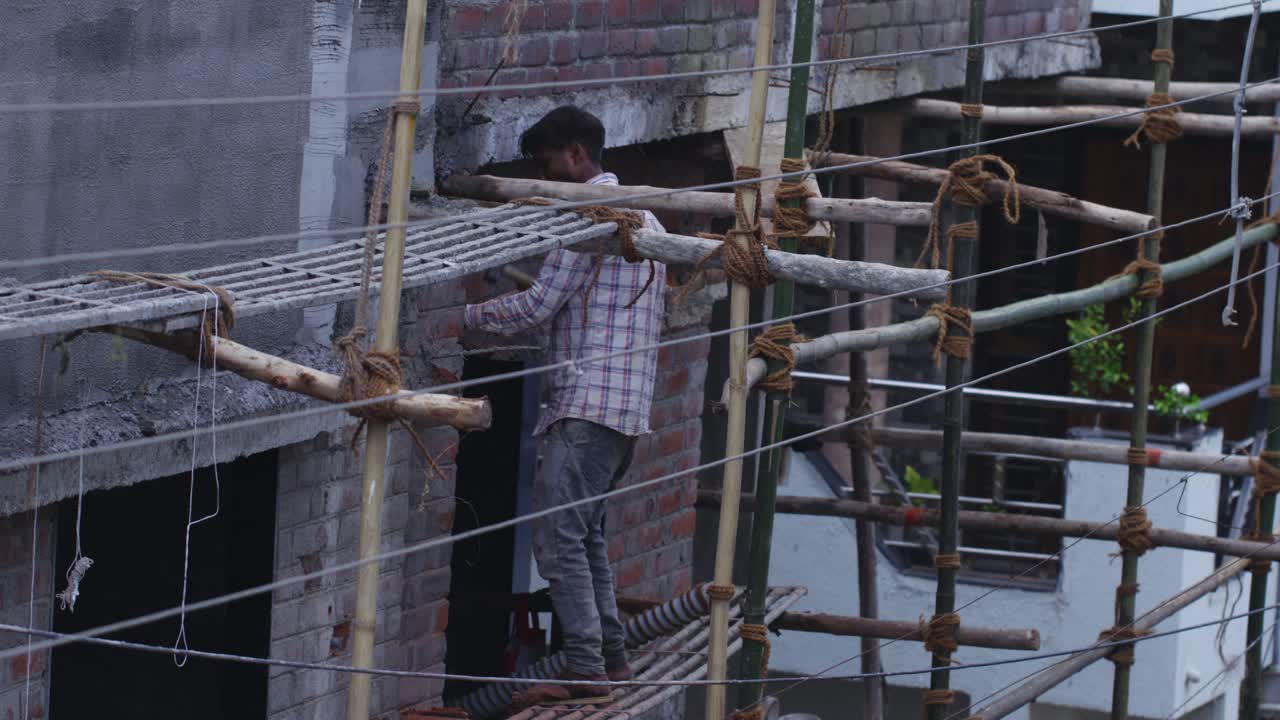A slow motion handheld long wide shot of an Indian man wearing checkered shirt, working at a construction site, hammering the wall, standing on a height amidst bamboo poles