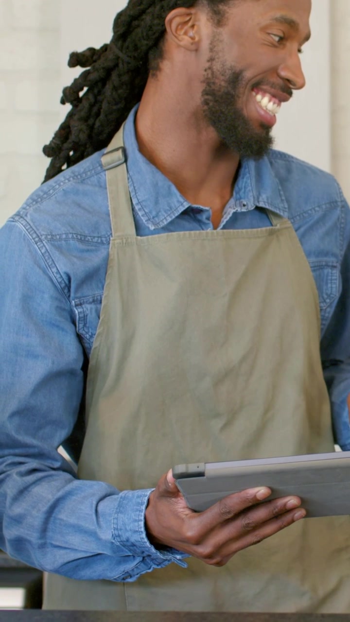 Vertical video: Helping offscreen guest, barista in olive apron tapping tablet, laughing at counter