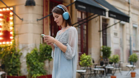 Happy-young-woman-in-wireless-headphones-choosing,-listening-music-in-smartphone-dancing-outdoors