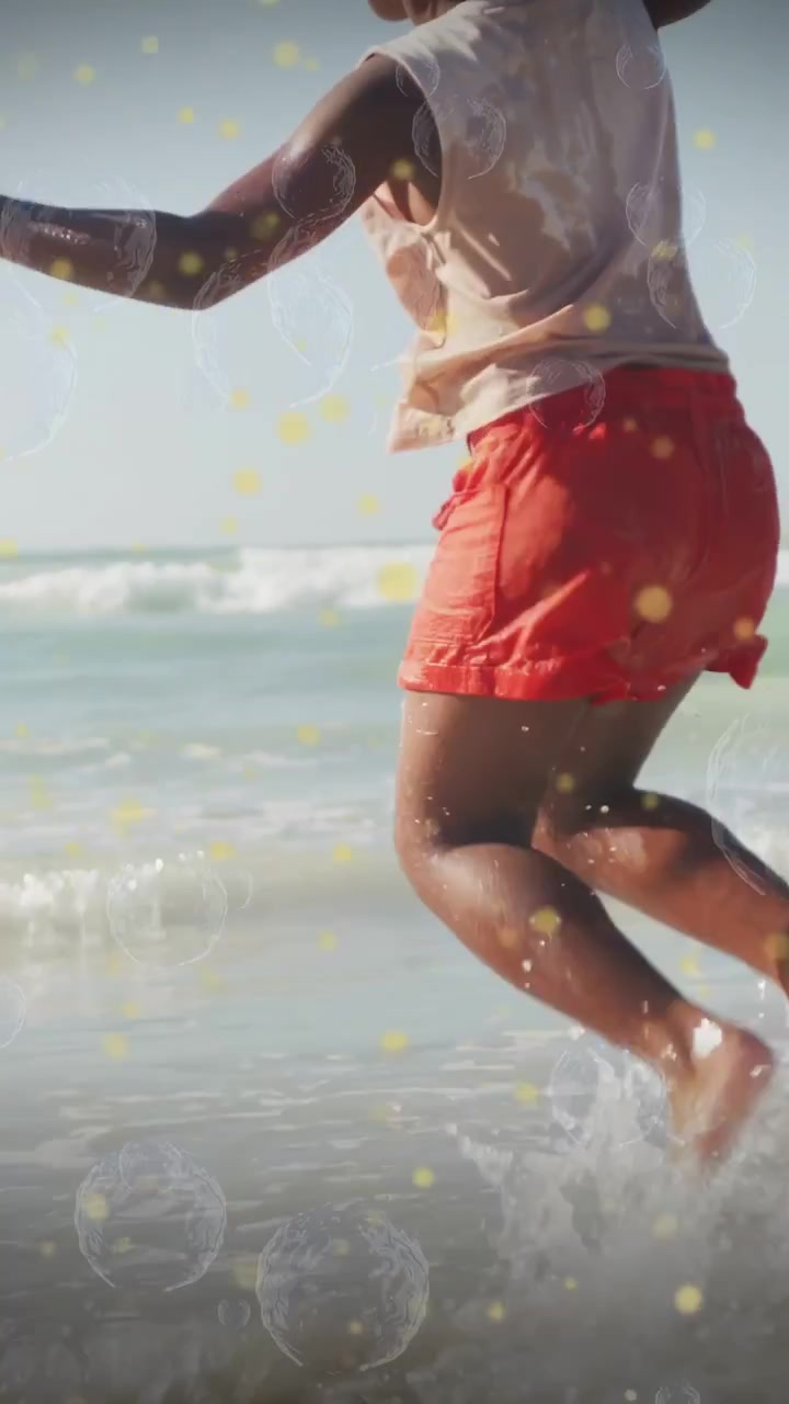Video verticale: Ragazza che salta sulla riva, un'onda che provoca uno schizzo, bolle con effetto bokeh che accentuano il gioco per il viaggio.