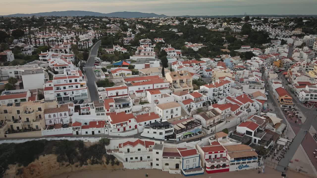 Camera glides above Carvoeiro’s rooftops and winding streets, moving inland from the golden beach to reveal the charming village architecture and patchwork of whitewashed facades