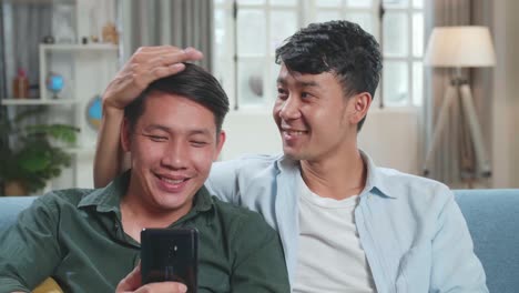 Asian Gentle Gay Couple Using Mobile Phone, While Sitting On A Couch In Cozy Stylish Apartment. Adult Boyfriends Online Shopping On Internet, Watching Funny Videos On Streaming Service