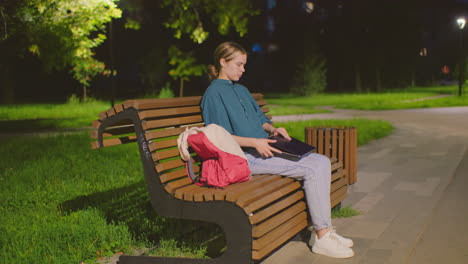 side view of young woman seated on outdoor bench at night, illuminated by soft lighting as she opens her laptop, red backpack rests beside her, surrounded by greenery and nighttime