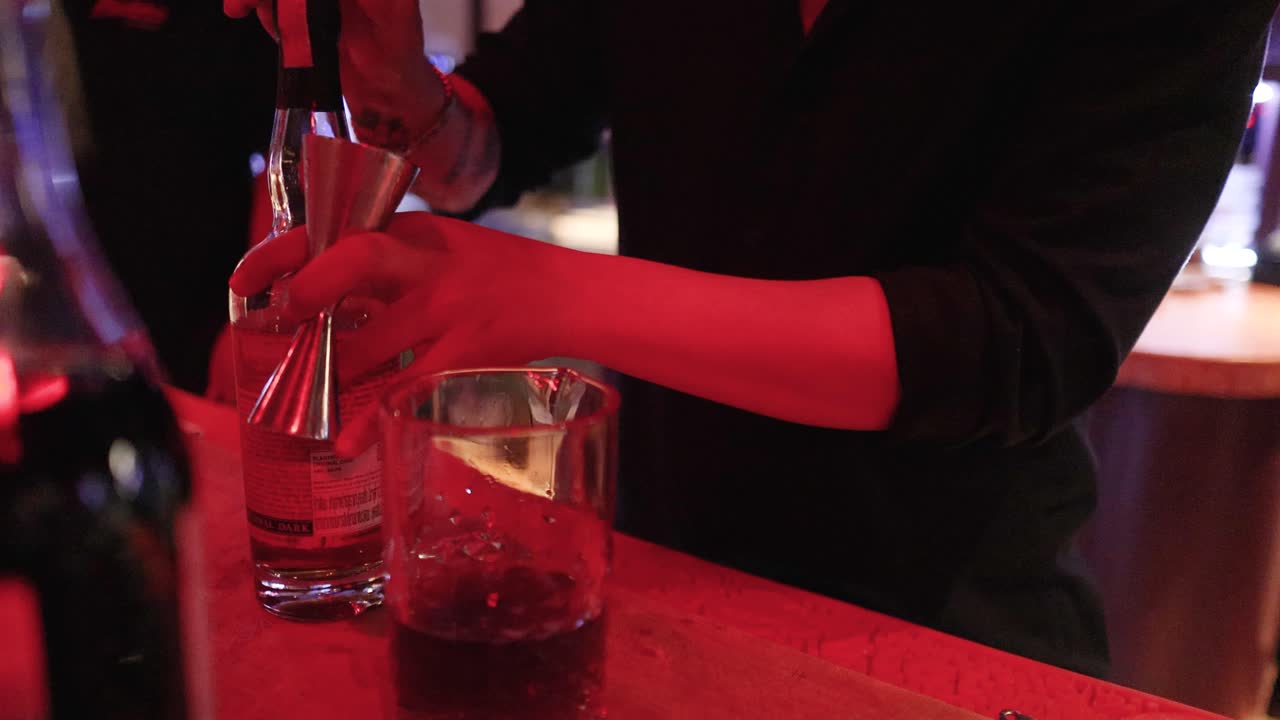 A bartender skillfully prepares a cocktail in a dimly lit bar, using precise techniques and tools under warm red lighting