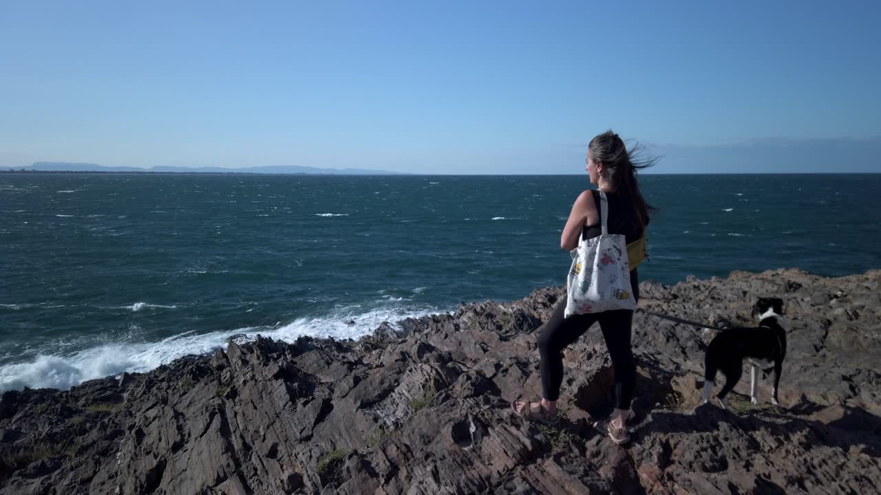 Female walking with her dog, wind blowing through her hair while standing on rocky mediterranean coastline in collioure, france, experiencing serene landscape in slow motion under sunny blue sky