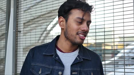 slightly angsty guy in jean jacket talks to someone out of frame and leans on the train station fence