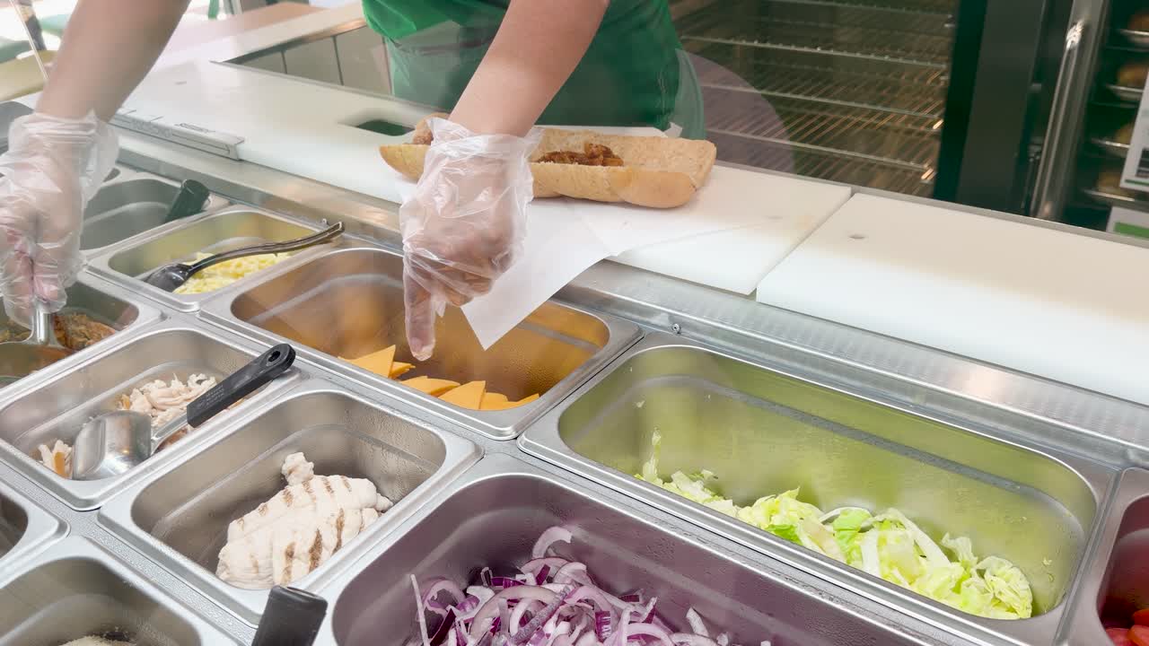 Un addetto con guanti protettivi aggiunge carne alla griglia a un panino in una cucina.