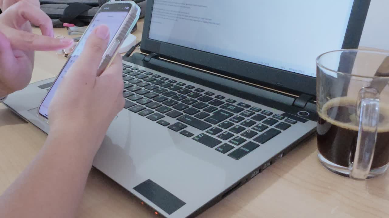 Home office desk setup: hands using smartphone with laptop, coffee, smartwatch, lamp, and stationery