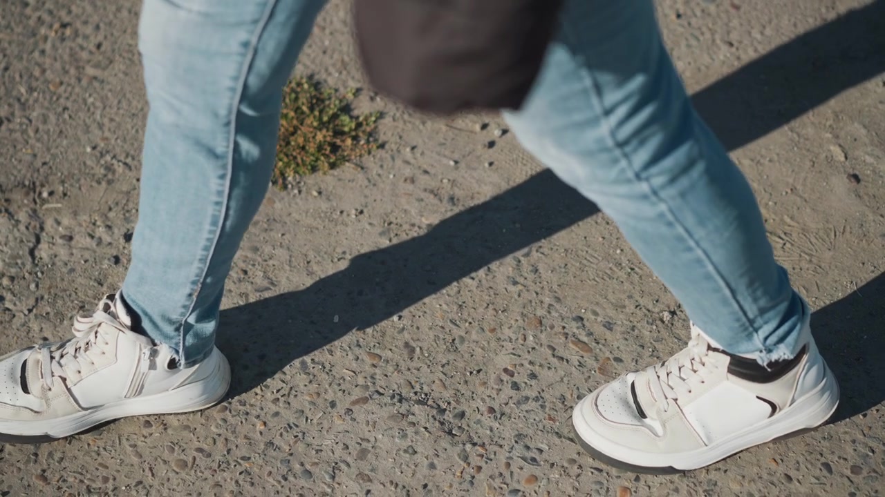 Una donna cammina su una superficie di cemento ruvida indossando jeans azzurri e scarpe da ginnastica bianche. L'inquadratura rimane costantemente dal basso sui suoi piedi mentre la donna avanza con la luce naturale del giorno che proietta.