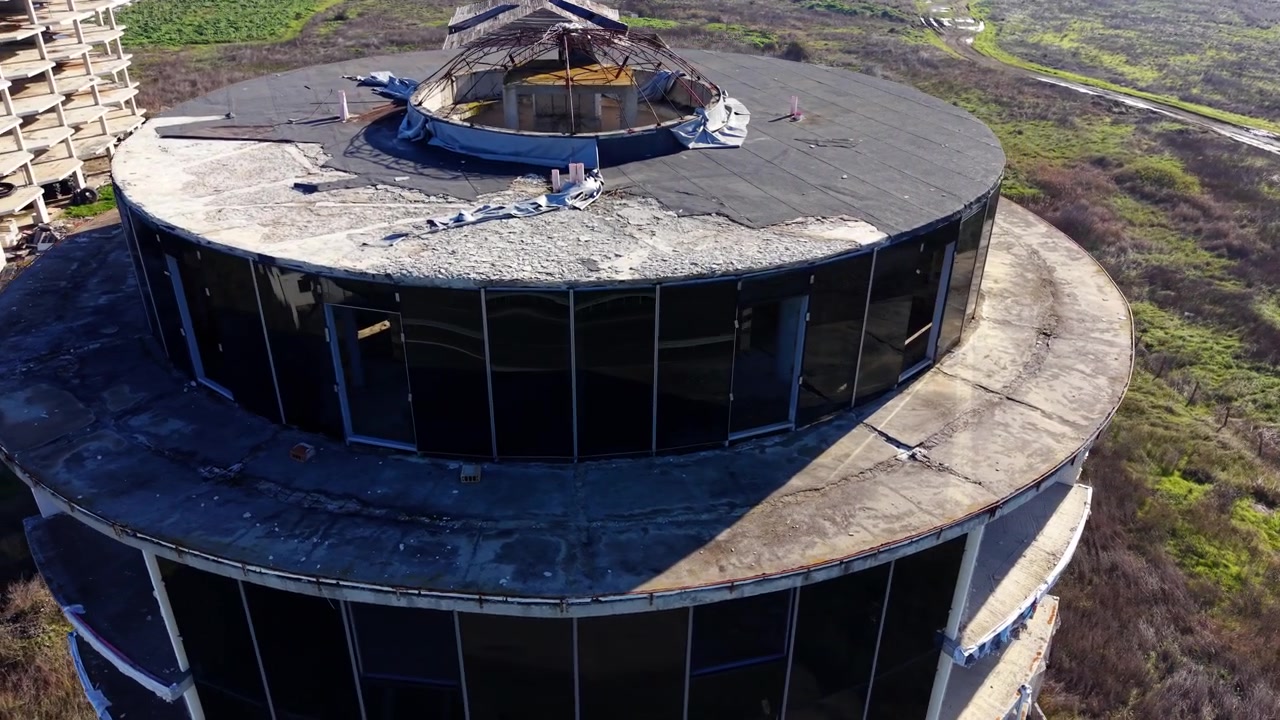 Vista aérea de un edificio redondo abandonado con techo de cúpula dañado. La vista de dron muestra una membrana rasgada, estructura expuesta, paredes de cristal y pastizales abiertos más allá.