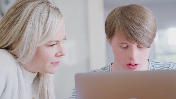 Mand med Downs syndrom sidder med kvindelig hjemmelærer og bruger bærbar computer til en lektion derhjemme.