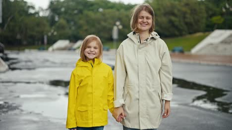 Porträt-Einer-Glücklichen-Frau-In-Einer-Weißen-Jacke-Und-Eines-Jungen-Mädchens-In-Einer-Gelben-Jacke,-Sie-Nehmen-Ihre-Kapuzen-Ab-Und-Posieren-Und-Lächeln-Nach-Dem-Regen-Im-Park