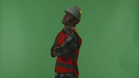 side view of asian male engineer with safety helmet crossing his arms and looking around while standing in the green screen background studio, during sunset or sunrise time