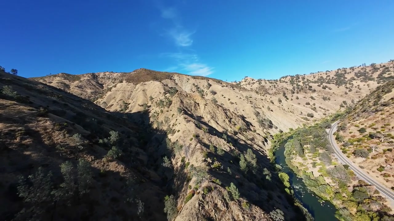 Vista aerea FPV di colline aride e ondulate nel nord della California, che rivela progressivamente le rigogliose sponde verdi di Cache Creek e una strada tortuosa incastonata nel canyon sottostante