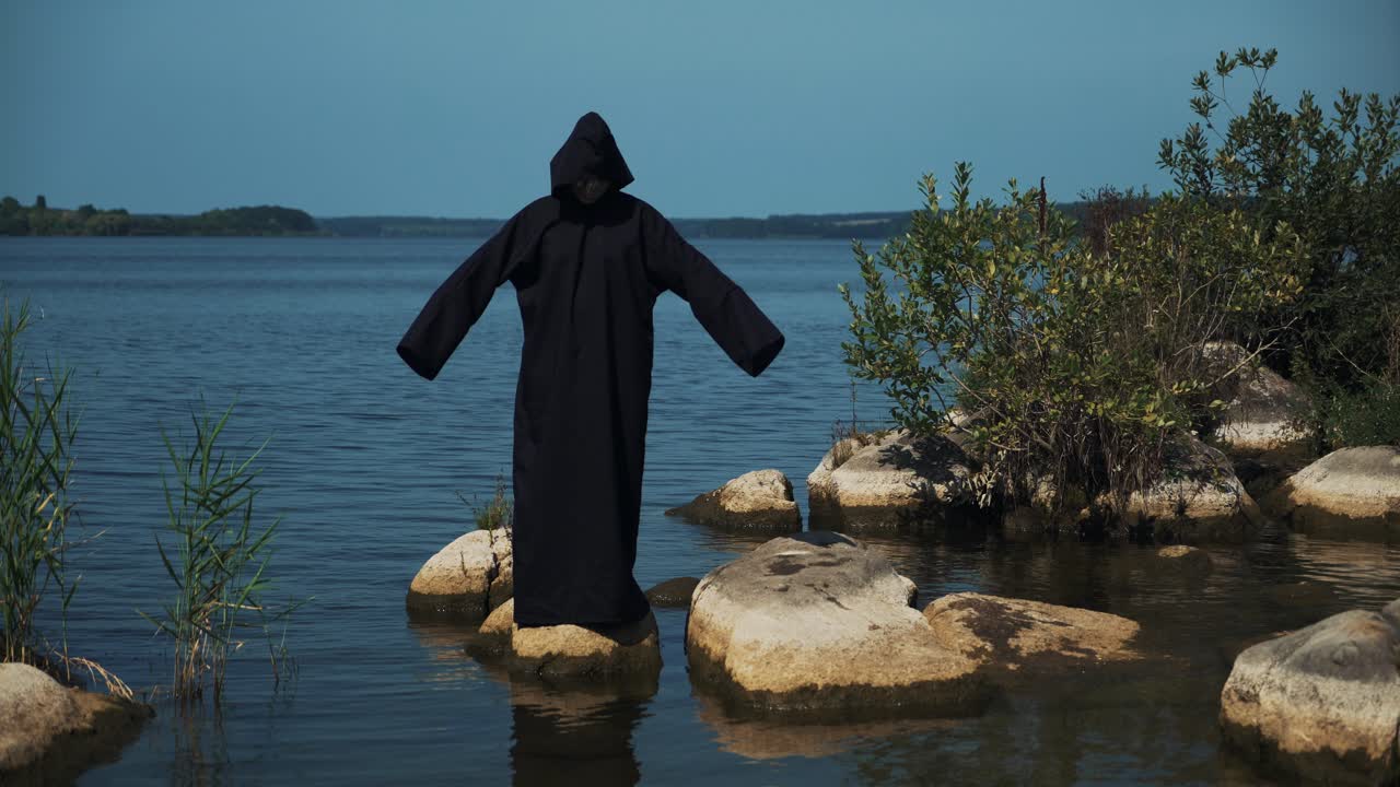 Woman devil in black cape and hood on stones near the river. Halloween costume.