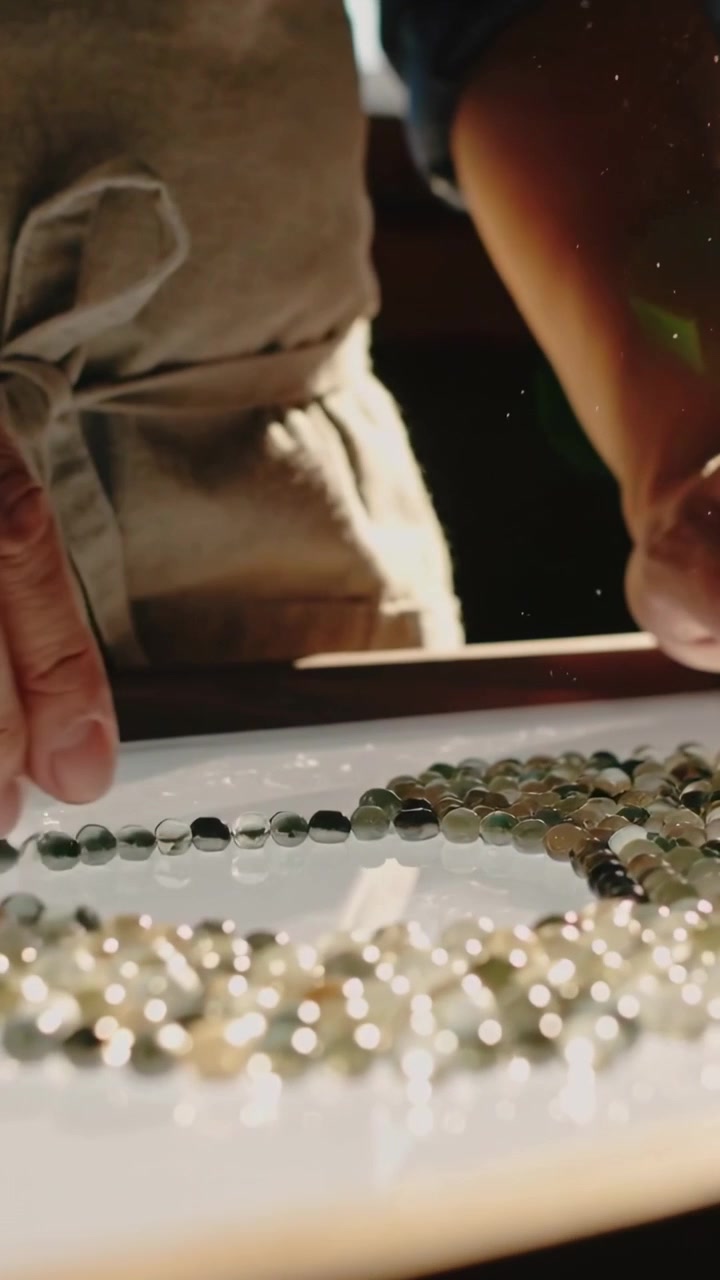 Vertical video: Sorting pro hands lining beads on lit tray by window, sun streaming, wearing apron