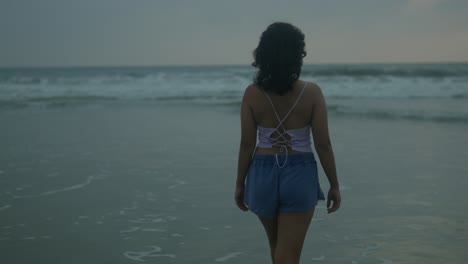 Young-woman-in-blue-shorts-standing-contemplatively-by-the-ocean-at-dusk,-gentle-waves-lapping-at-the-shore