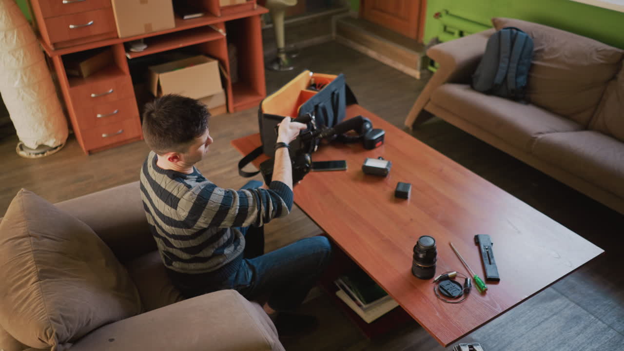 Young man organizes camera equipment, adjusting microphone and placing gear on table in cozy home studio. Work desk cluttered with lenses, batteries, and other filming tools