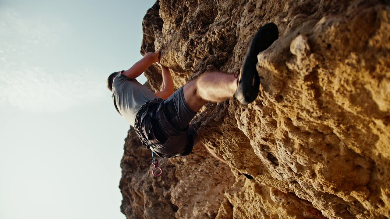 desde abajo, un hombre con una camiseta gris y pantalones grises en un seguro especial sube una roca empinada amarilla y piedras contra el telón de fondo de un cielo soleado y despejado de verano