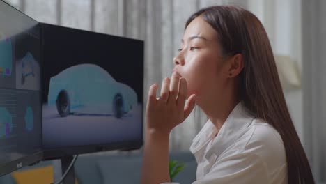 close up side view of asian female automotive designer holding hands on cheek while working boringly on 3d model of ev car on the desktop computers in the studio