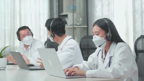 a woman of three asian doctors with stethoscope in headset and mask working as call center agents speaking on a call and typing while her colleagues are talking during working at the office