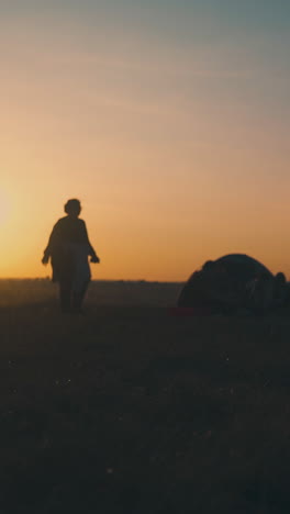 silhouette of overweight woman dancing near camping friends at bonfire on river bank against setting sun low angle shot