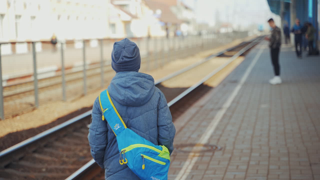 Young boy in winter clothes waiting for the train on the background of a railway station. Back view of one boy with a bag on his back standing on the platform and looking ahead.