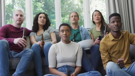 group of diverse young people holding beers cheering while watch tv at home