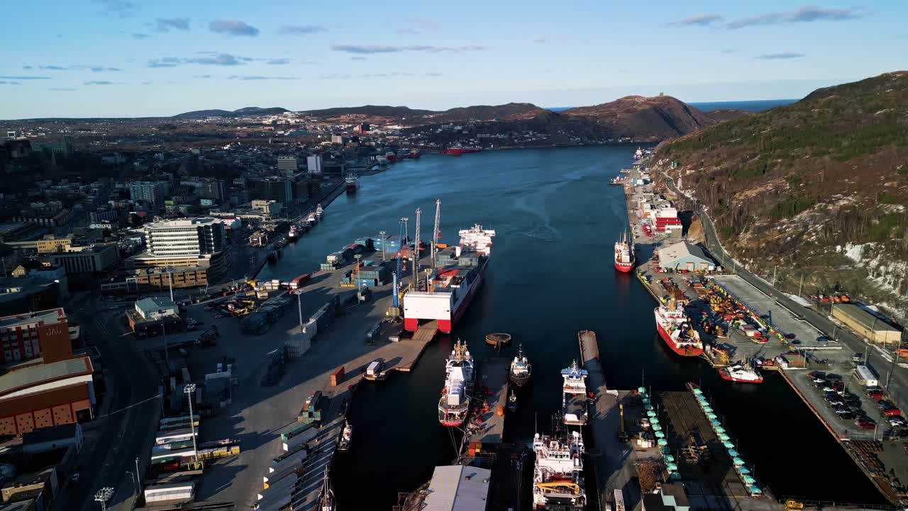A sweeping drone shot looks down the length of St. John's harbour in Newfoundland, showing downtown buildings on one side, ships and docks on the other, and hills rising above the Atlantic