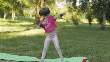 athletic child girl in vr headset helmet making fitness workout exercises in park. virtual reality