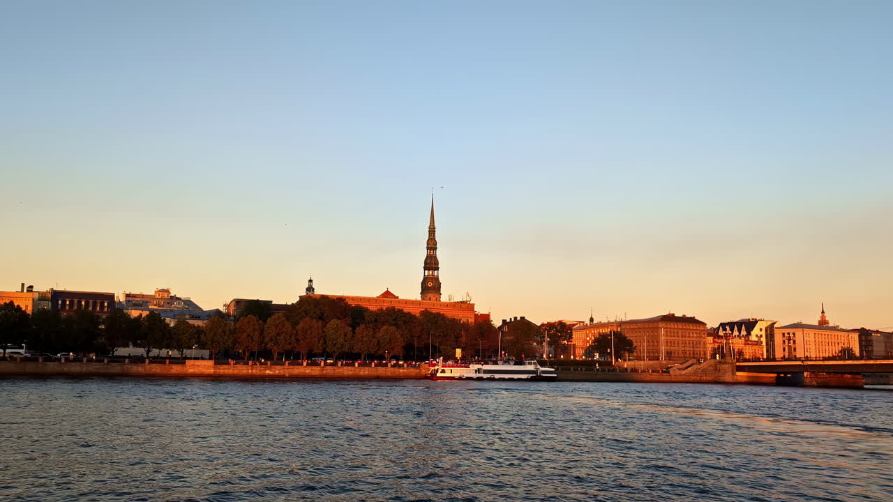 Skyline di Riga al tramonto con riflessi sul fiume, edifici storici e una calda luce dorata serale.