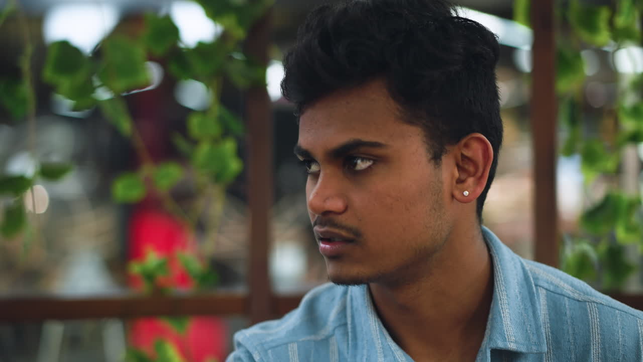 Indian man focused on conversation while eating potato chips, closeup showing attentive expression, hand holding crinkled chip bag, indoor setting with blurred background, subtle smile and eye contact