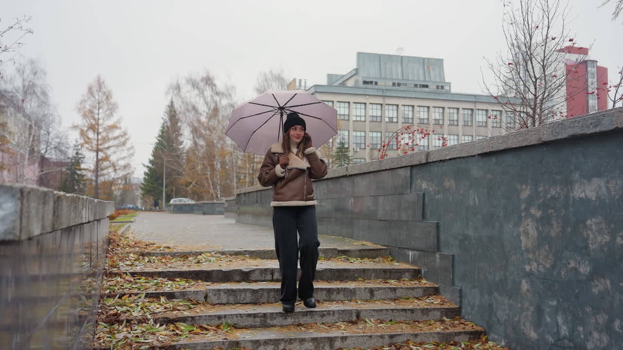 Student smiling on phone call holding umbrella, wearing knit cap, brown shearling jacket, black trousers, walking down stone steps during light snowfall with colorful autumn leaves