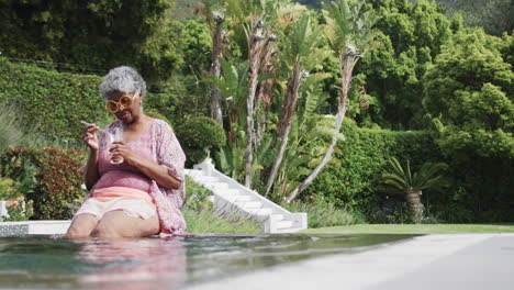 Feliz-Mujer-Afroamericana-Mayor-Sentada-Con-Piernas-En-La-Piscina-Sosteniendo-Una-Bebida,-Espacio-Para-Copiar,-Cámara-Lenta