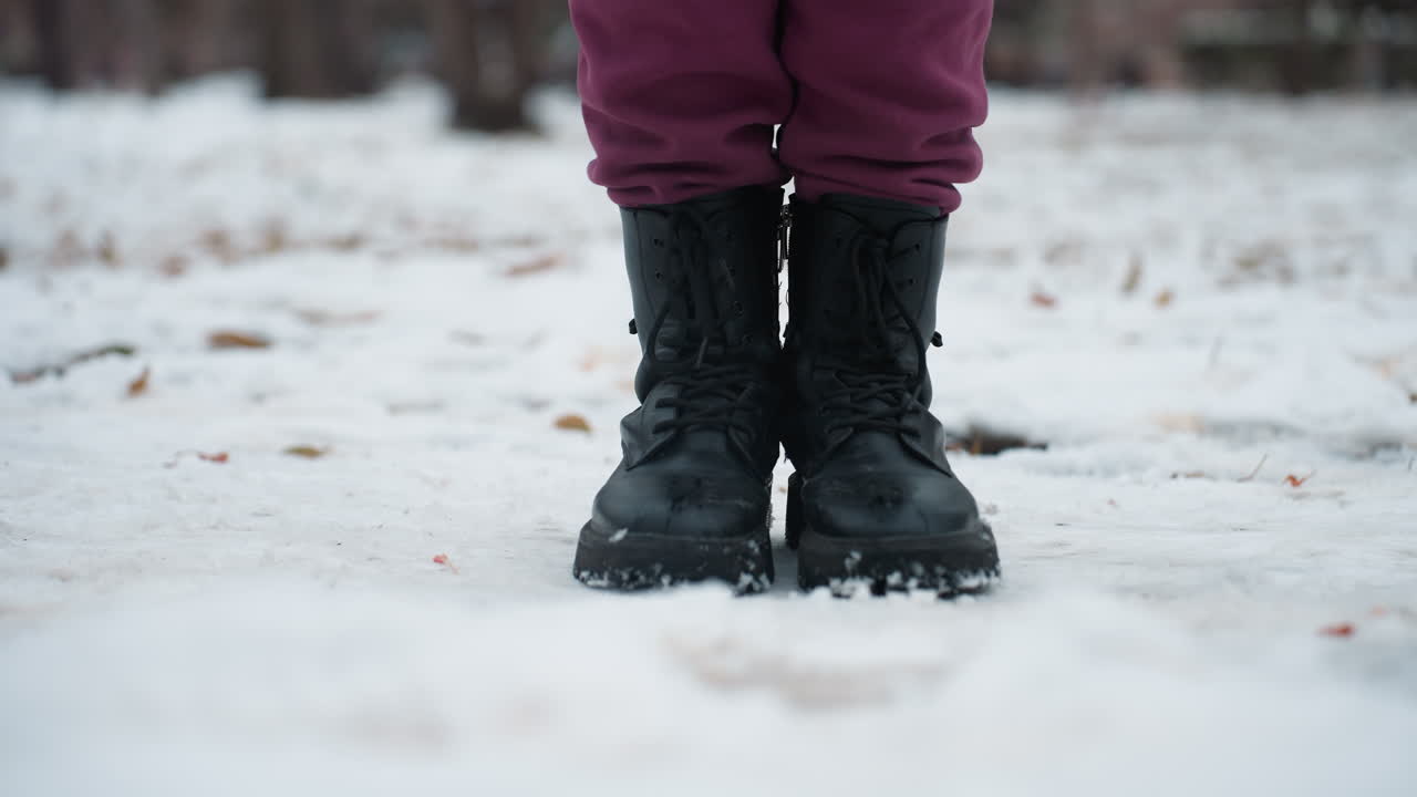 primer plano de piernas con botas negras mientras la persona salta de izquierda a derecha en el suelo cubierto de nieve, las botas dejan ligeras huellas en la nieve fresca, con aire frío de invierno y fondo borroso