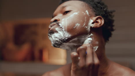 black man carefully shaving face with razor in front of bathroom mirror