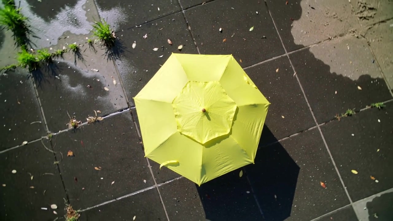 La telecamera si allontana rivelando un ombrellone giallo acceso su un terrazzo piastrellato, mostrando pozzanghere e ciuffi d'erba.