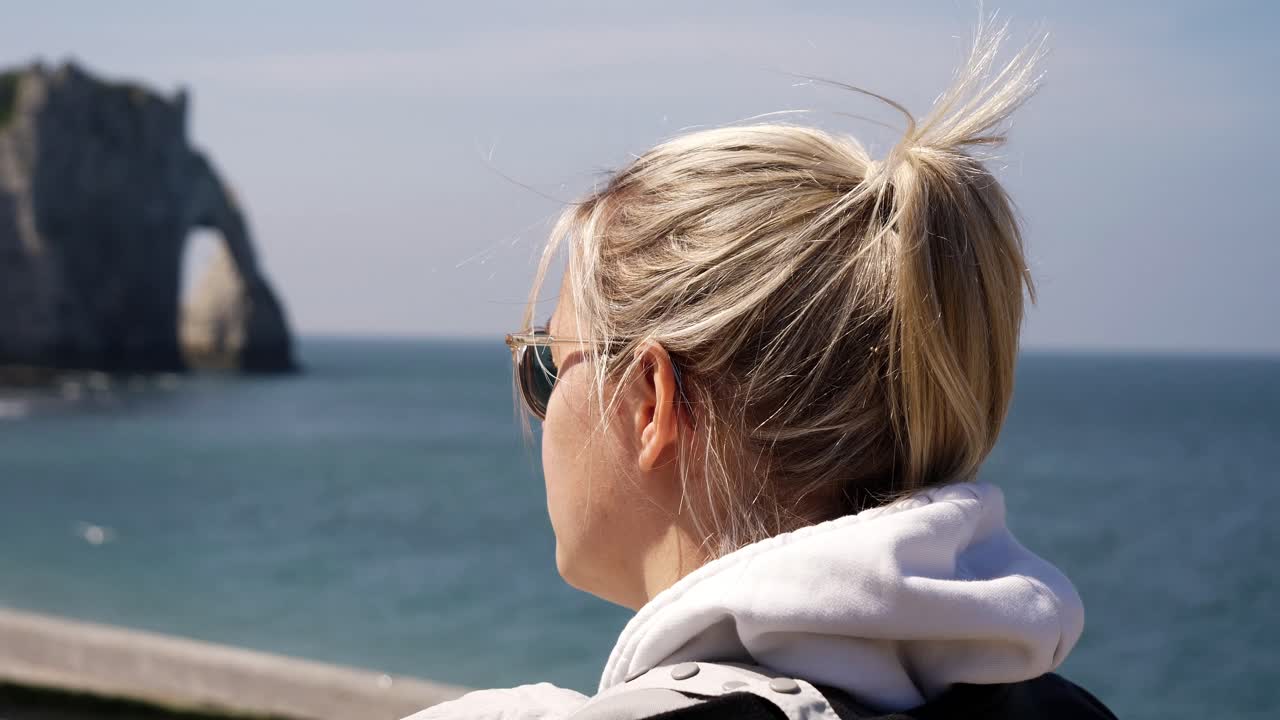 Slow motion close up of blond woman walking along promenade of Etretat with famous sea cliffs in background