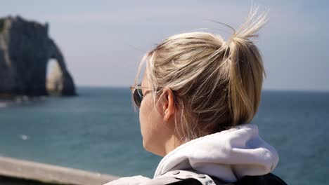 Slow-motion-close-up-of-blond-woman-walking-along-promenade-of-Etretat-with-famous-sea-cliffs-in-background-