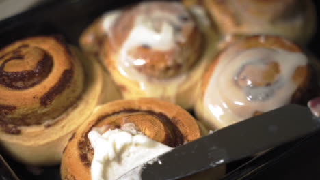 close up knife spreading sweet icing on warm homemade cinnamon buns