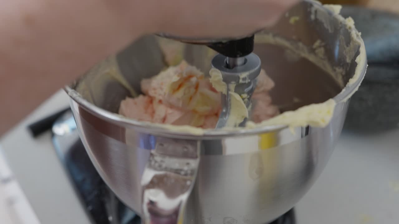 Close up, A cook taps a spatula on the side of a mixer bowl to tap off excess buttercream icing, then lowers the paddle and puts the mixer on low speed to whip the icing