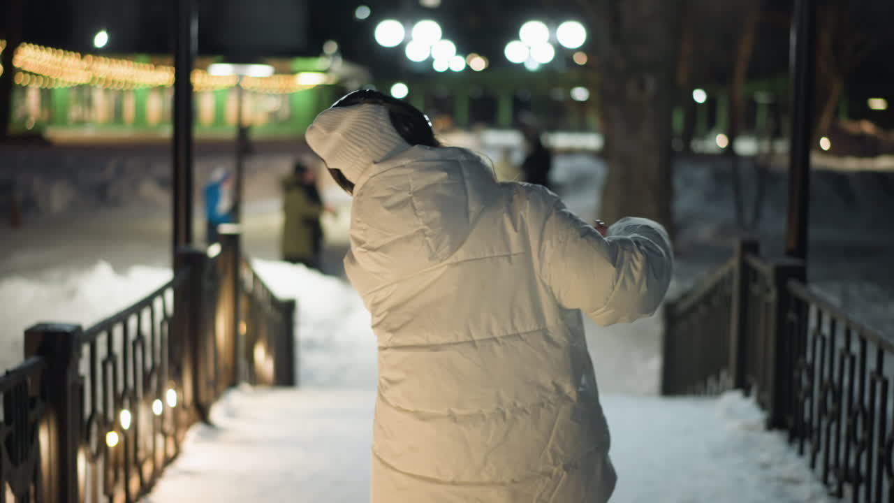Back view of girl spinning on snowy square under street lights at night, joyful expression, flowing arms in white coat and headphones, ambient urban park scene with people in background