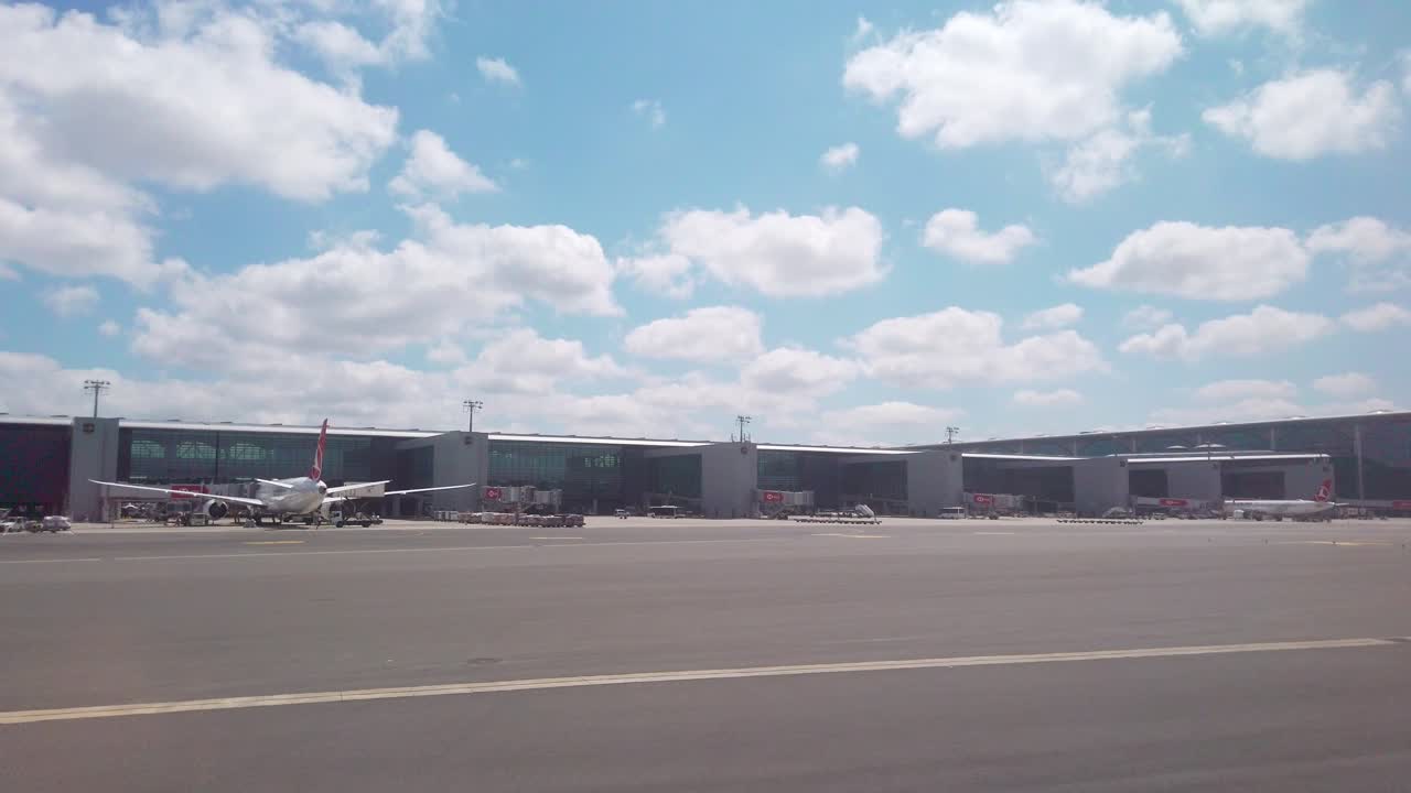 On a partly cloudy daytime, the view from the airplane window captures Istanbul Airport's terminal building and parked aircraft on the apron