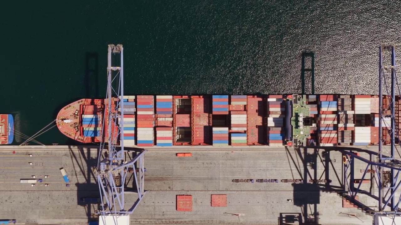 Pemandangan udara dari atas derek gantry yang memuat dan membongkar kontainer kargo warna-warni di kapal besar yang berlabuh di pelabuhan pengiriman komersial, mengilustrasikan perdagangan dan logistik global