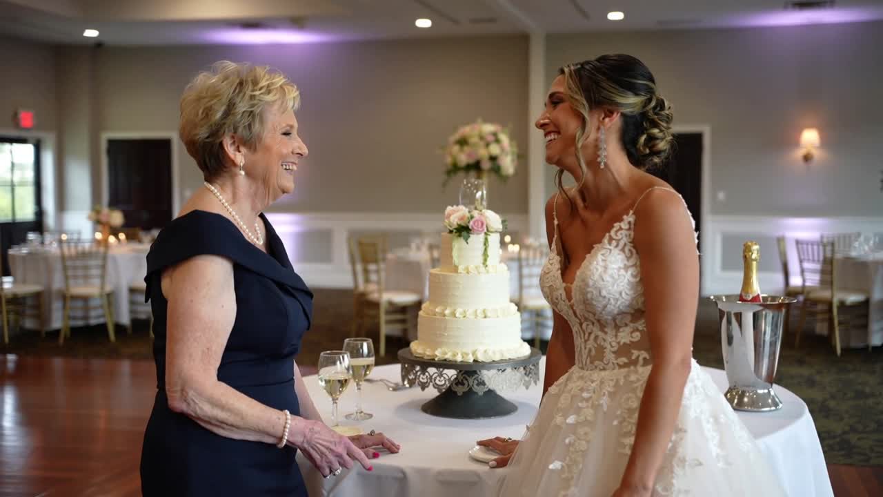 Sposa in abito che si appoggia e posa la mano su un tavolo al ricevimento, mentre un parente inizia una conversazione accanto alla torta.