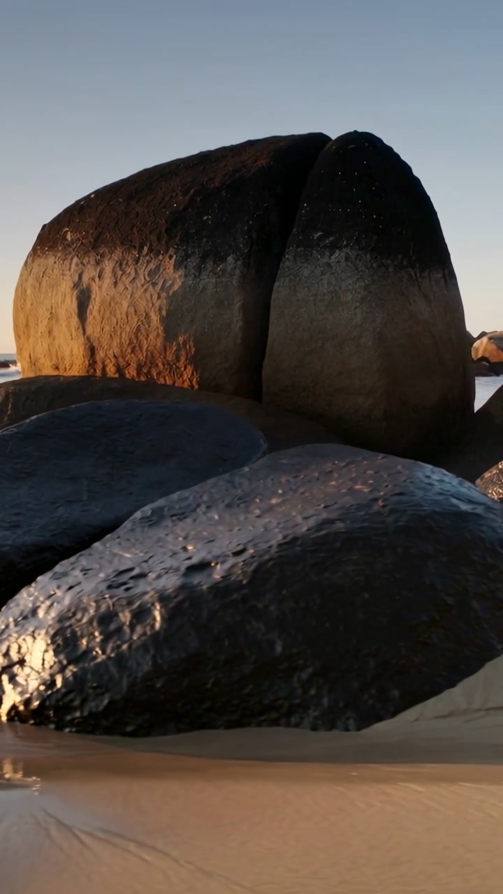 Vertical video: Camera pulling back, revealing split boulders on wet sand, showing sunset light