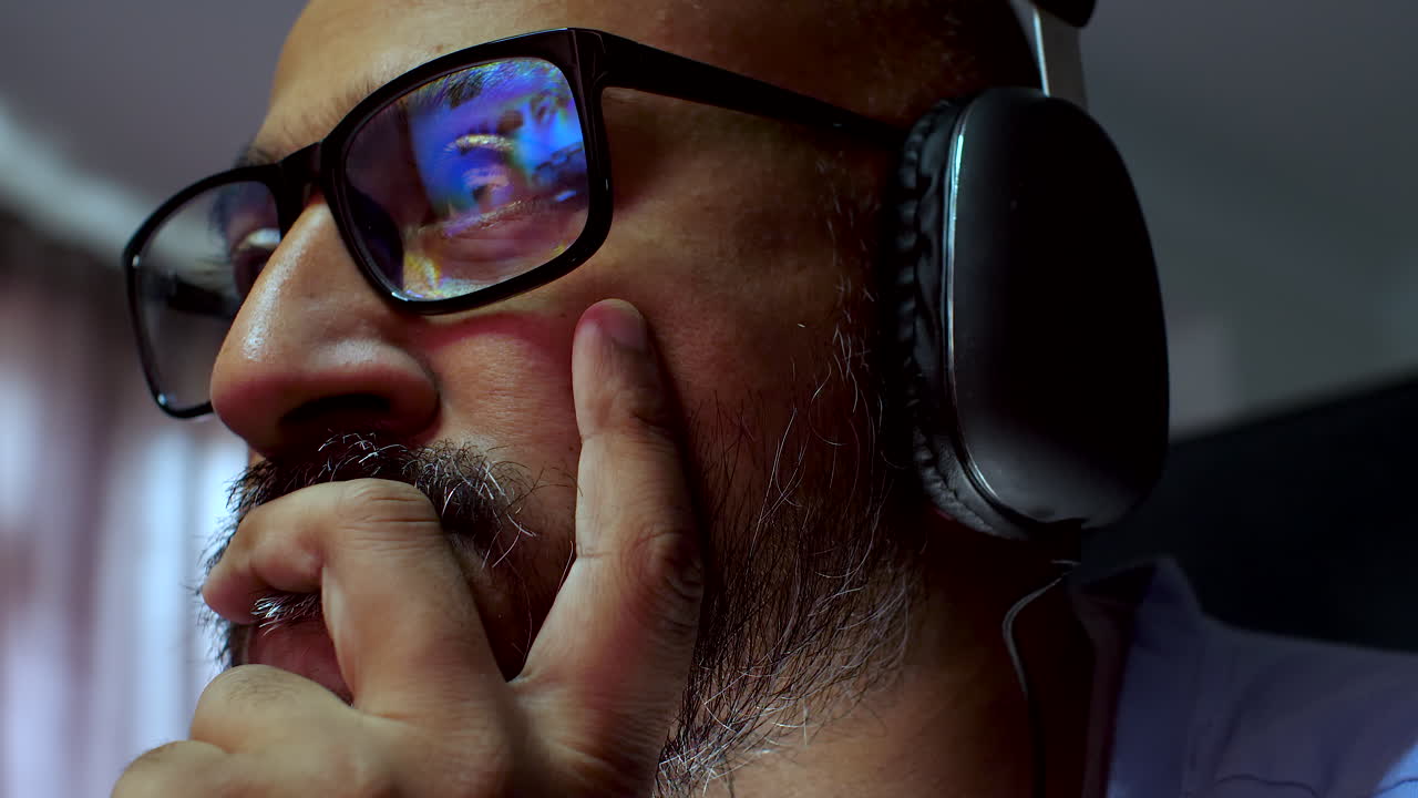 A close-up side profile view of a person with a beard wearing black over-ear headphones, with partial eyeglasses visible, creating a focused and immersive atmosphere
