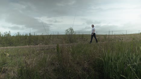 a man dressed in a white t-shirt and dark pants strolls along a dirt road surrounded by expansive green fields