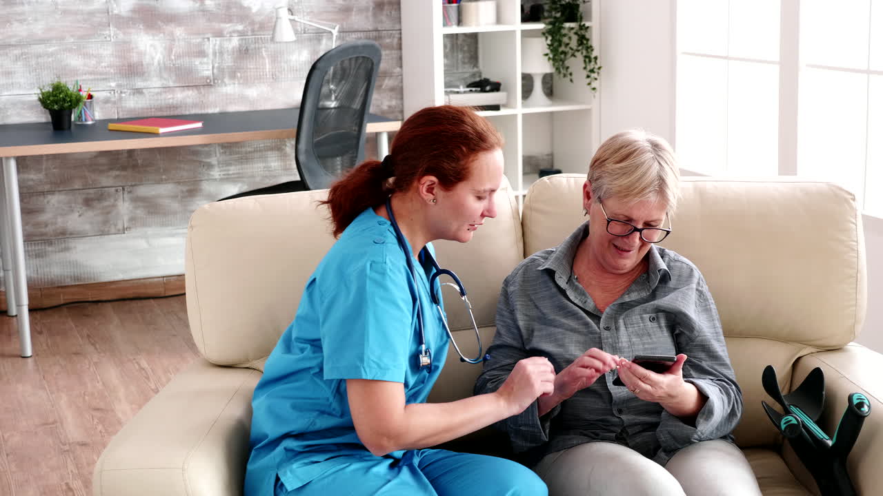 Nurse in retirement home sitting on the couch helping elderly woman to use a smartphone