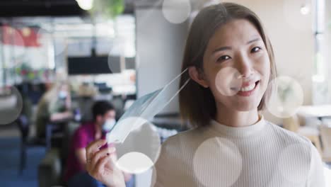 animation of light spots over asian businesswoman taking face mask off and smiling